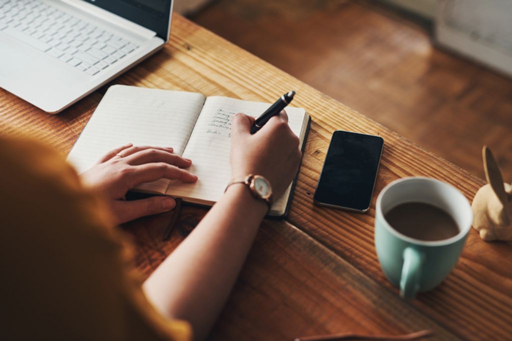 Shot of a woman's hand writing new year's resolutions in a notebook on a desk with a cup of coffee
